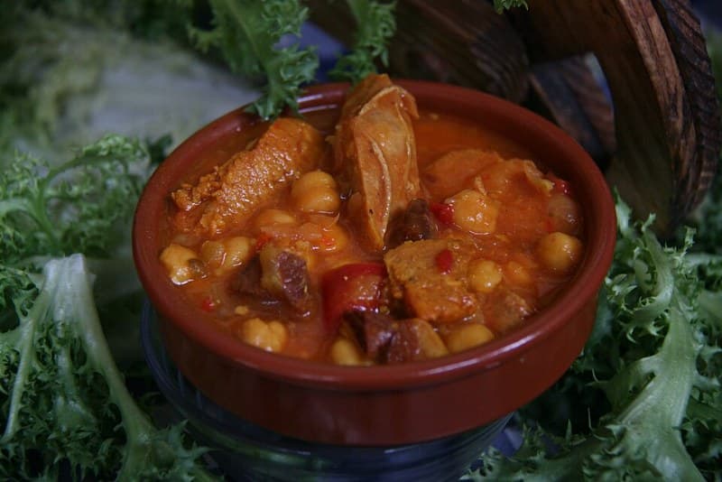 Clay bowl of hearty stew with chickpeas, sausage, meat, and vegetables.