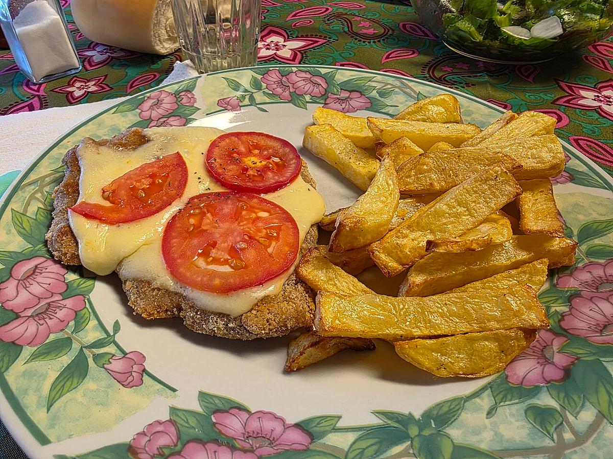 Escalope panée garnie de fromage fondu et de tranches de tomate, servie avec des frites.