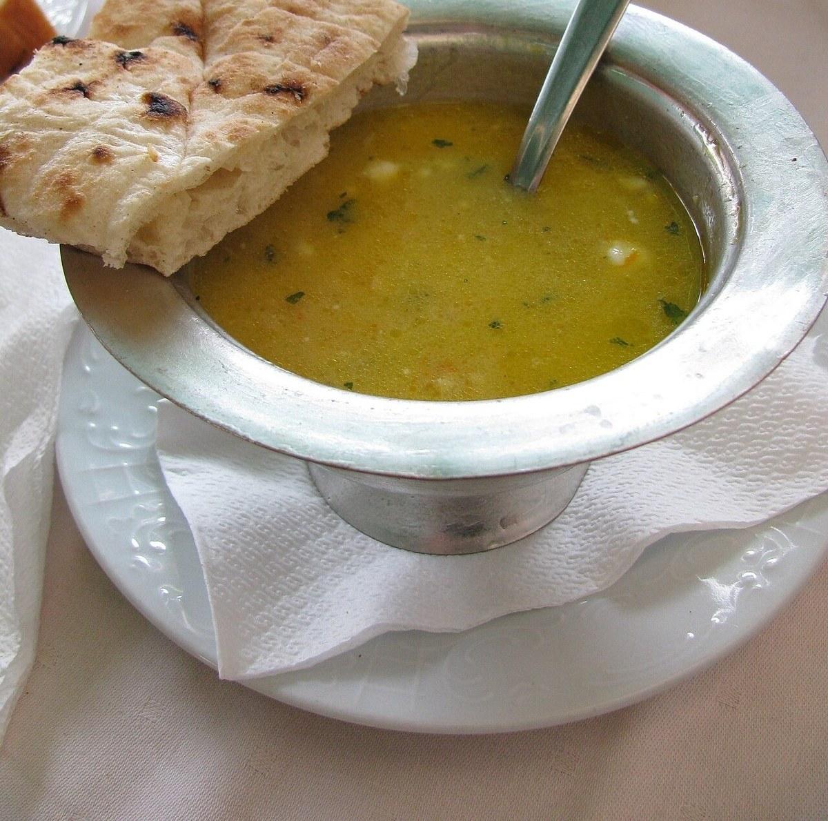 Metallschale mit gelber Huhn-Okra-Suppe, Fladenbrot und Löffel.
