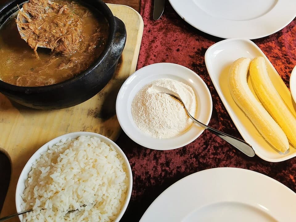 Table setting with a black pot of shredded beef stew, rice, banana pieces, and cassava flour.