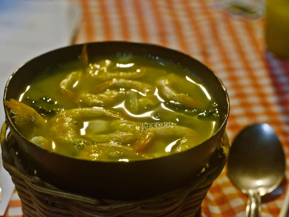 深色碗中黄色汤汁，含绿叶和虾，旁有勺子置于桌上。