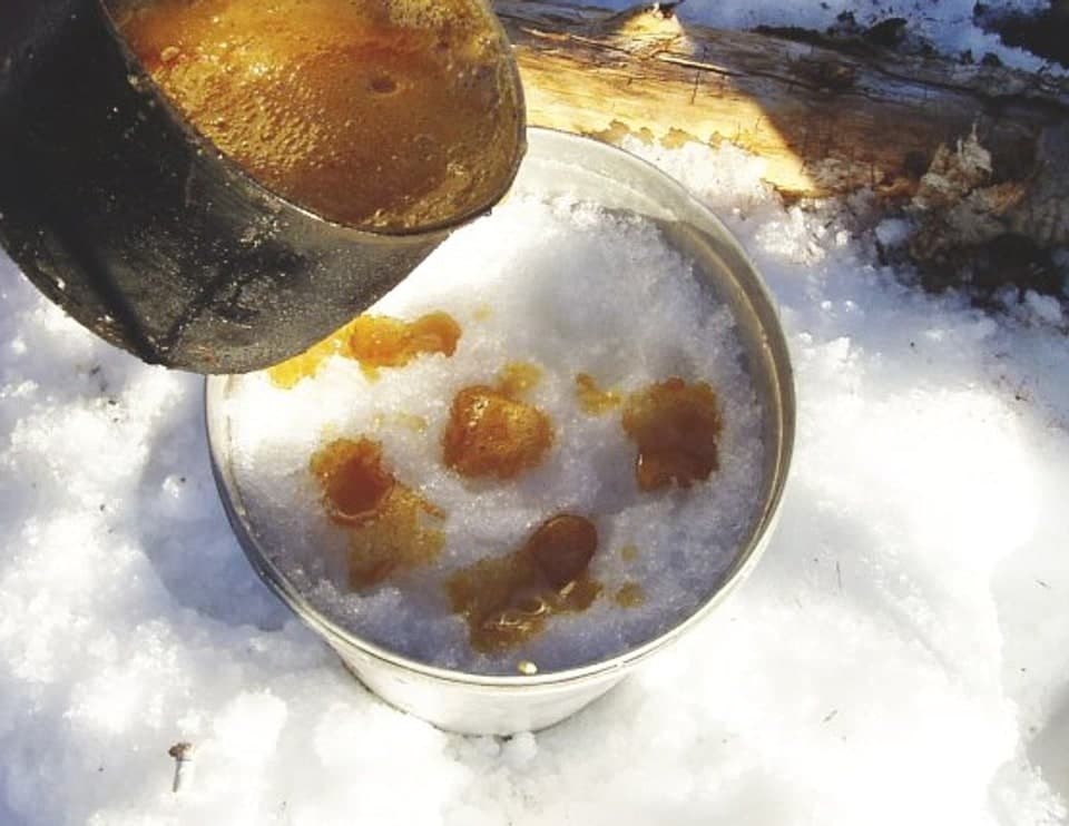 Bernsteinfarbener Ahornsirup, der auf Schnee in Patches abkühlt, neben einer Metallpfanne.