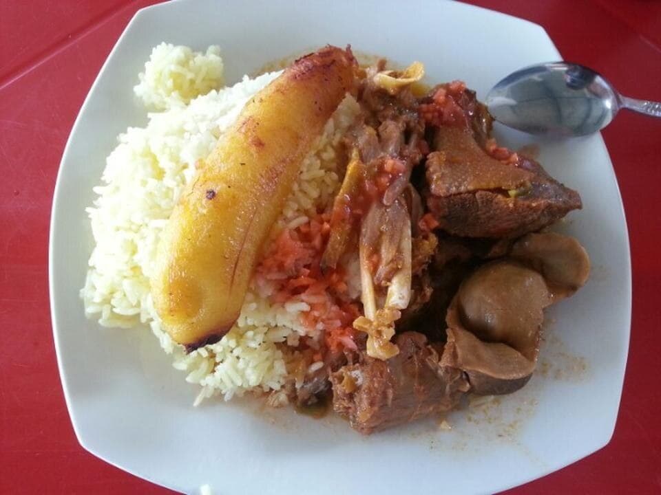 Ensopado de cabra em molho marrom servido com arroz branco e banana-da-terra madura frita em um prato branco.