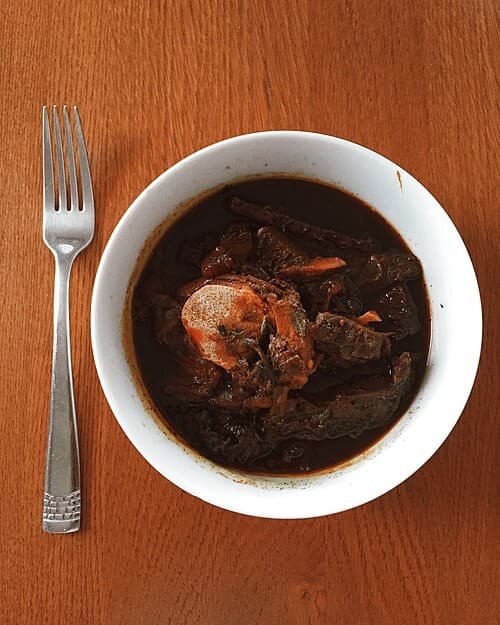 Tigela de ensopado escuro de carne sobre uma mesa de madeira com um garfo ao lado.