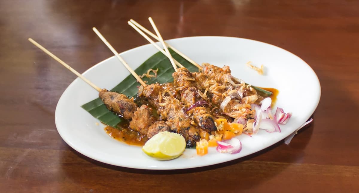 Pinchos de pollo en un plato con salsa de maní, lima, chalotas y pequeños acompañamientos.