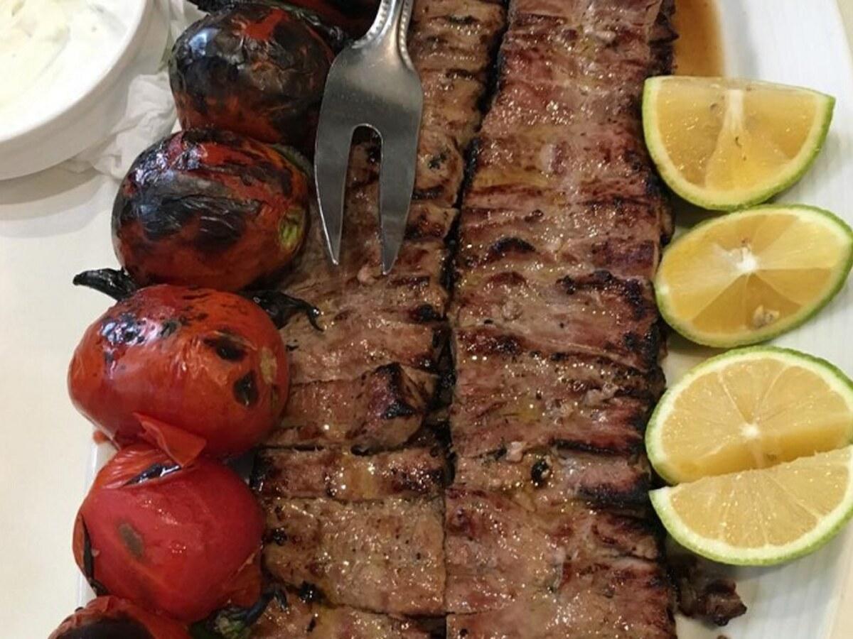 Grilled meat skewers beside charred tomatoes and lemon wedges on a plate.