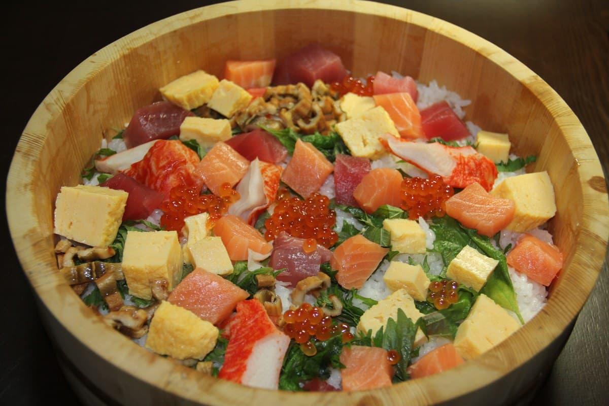 Wooden bowl of vinegared rice topped with raw fish, egg, vegetables, and garnishes.