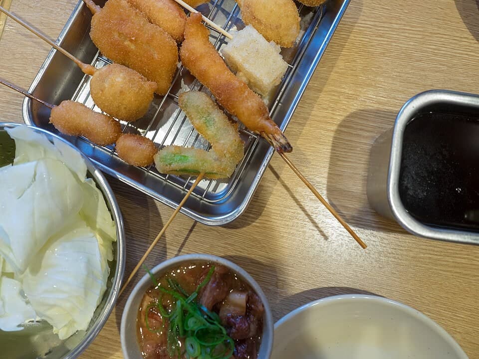 금빛 튀김 꼬치들이 금속 바구니에 담기고, 딥핑 소스가 가까이 놓여 있음.