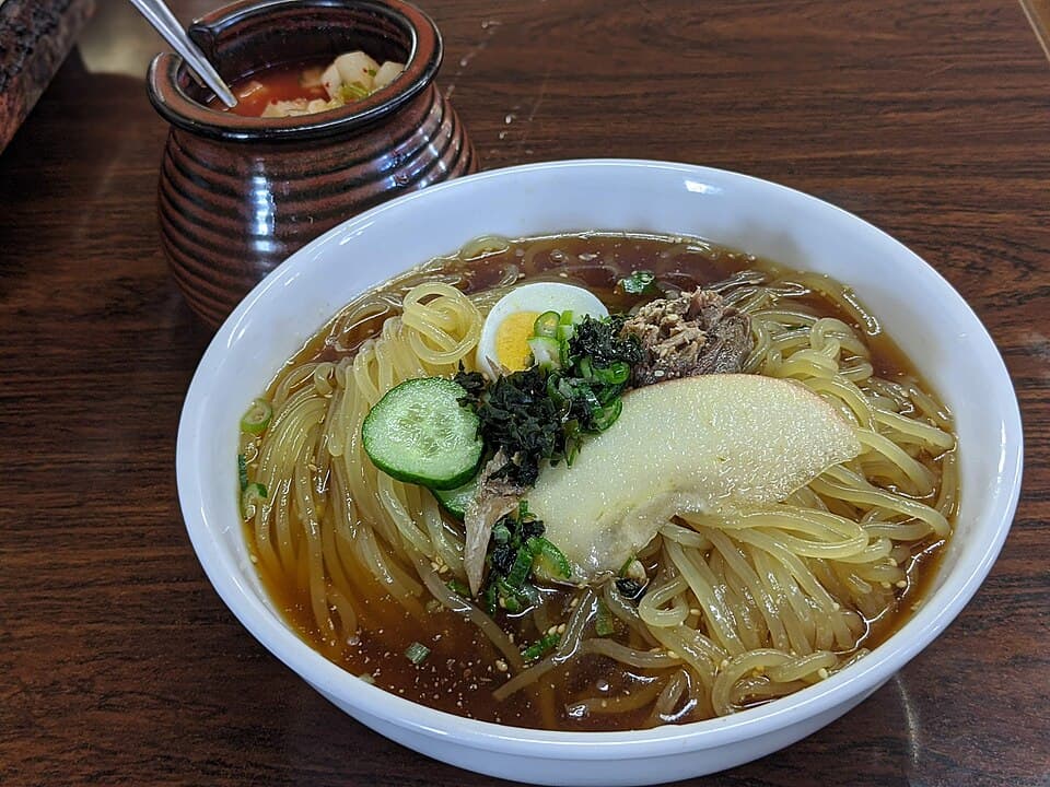 Un tazón de fideos fríos en caldo claro con pepino, huevo, kimchi y una guarnición de fruta pálida.