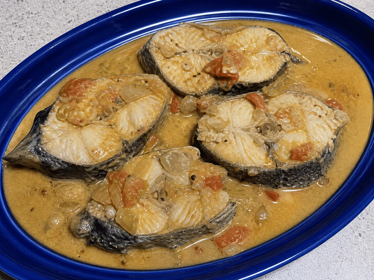 Filetes de pescado con piel en salsa de coco tostado con tomate y cebolla en una bandeja azul.
