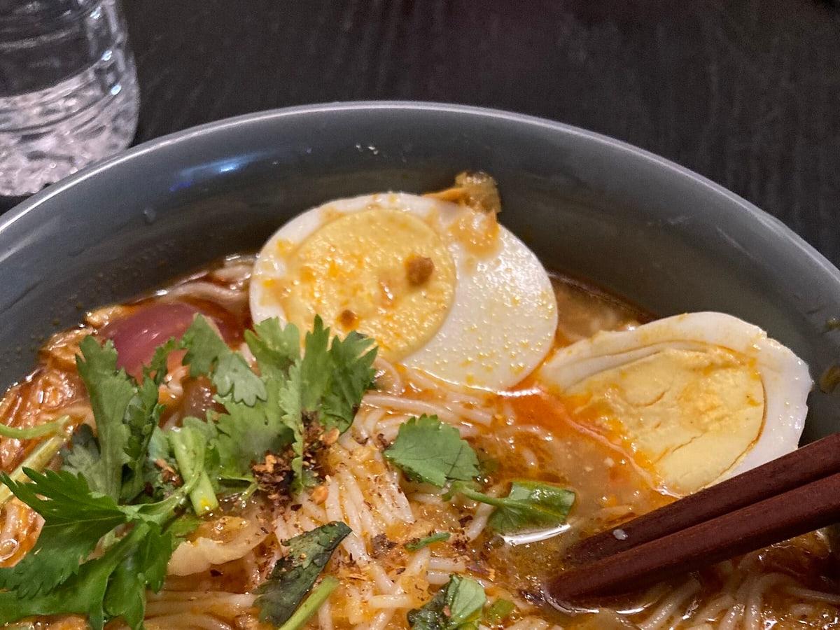 Bowl of rice noodle fish soup with egg, herbs, crispy toppings, and chopsticks.
