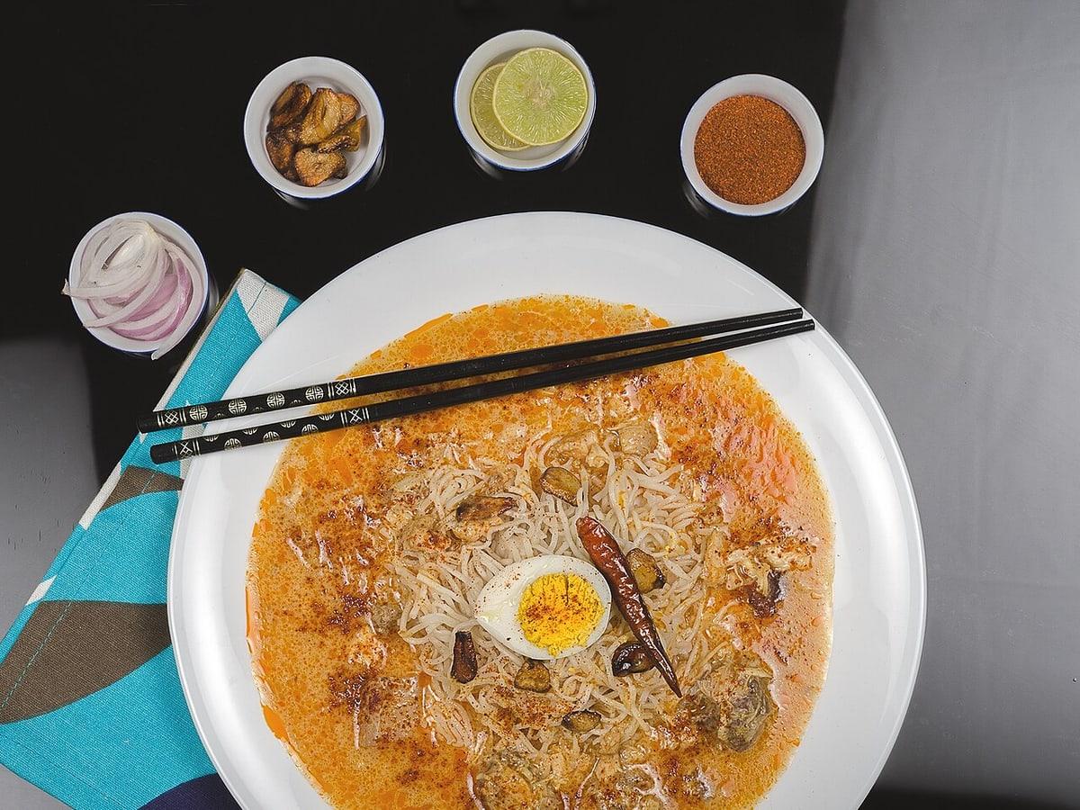 Schüssel Kokosnuss-Hühnernudelsuppe mit Ei, Nudeln, Limette, Zwiebeln und kleinen Gewürzschalen.