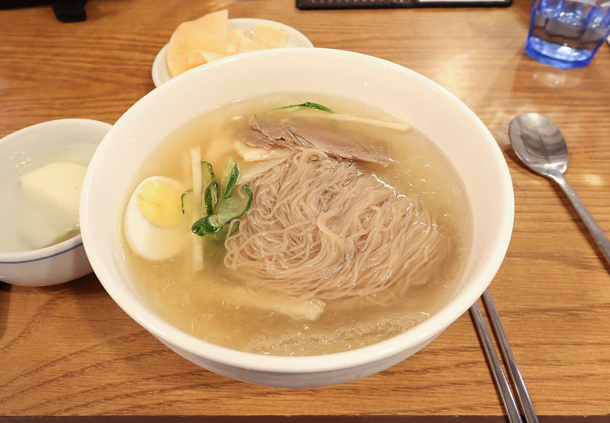 Bowl of cold buckwheat noodles in clear broth with egg, cucumber, and sliced meat.