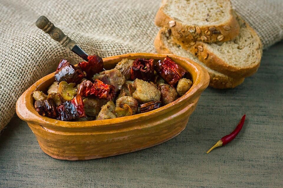 Clay pot filled with pork, mushrooms, onions, and dried peppers beside bread.