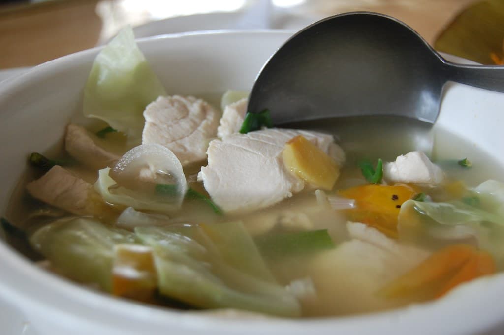 Sopa de pescado clara con jengibre, verduras y una cuchara en un recipiente blanco.
