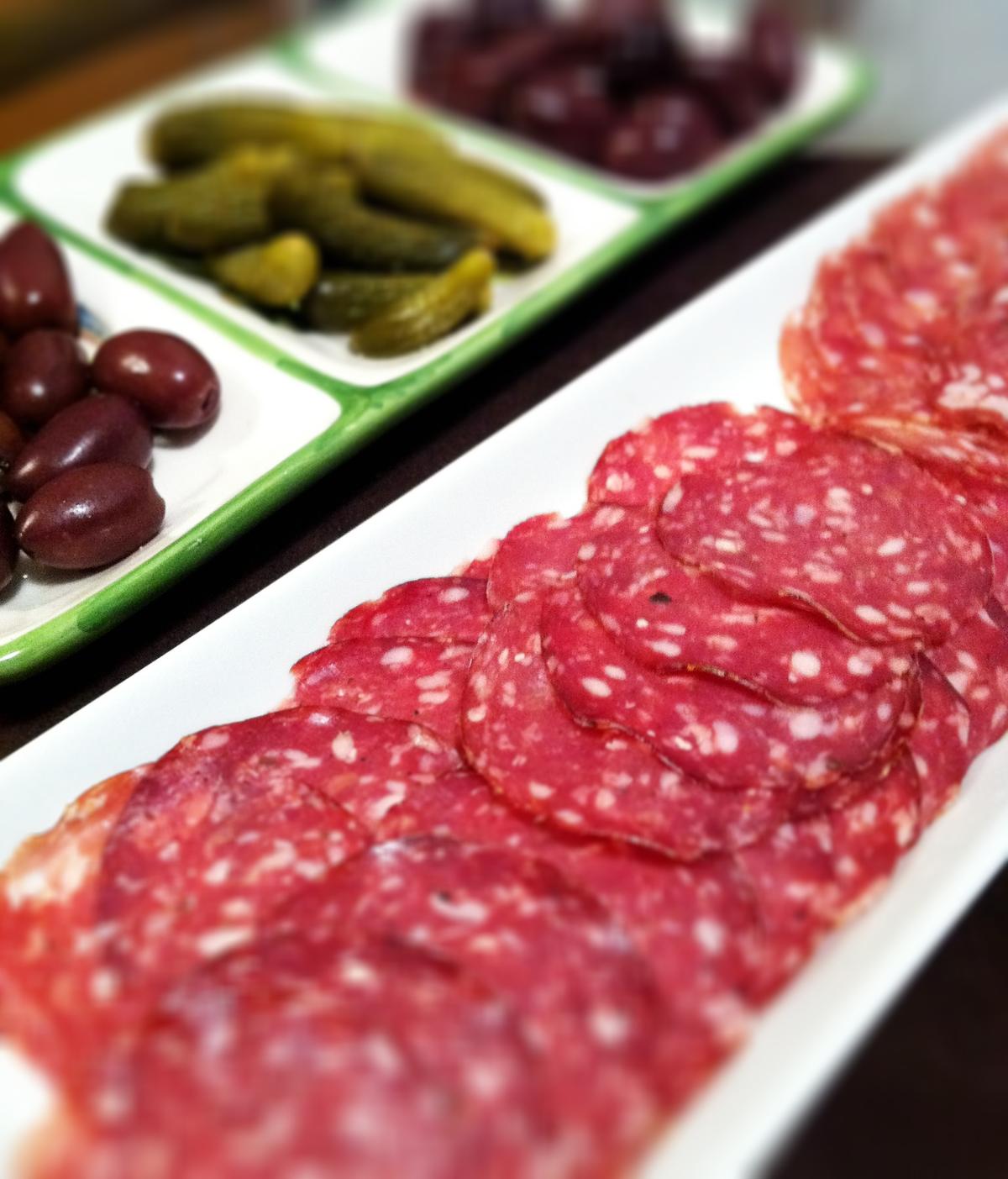 Fatias finas de salsicha curada vermelha servidas em um prato branco com azeitonas e picles.