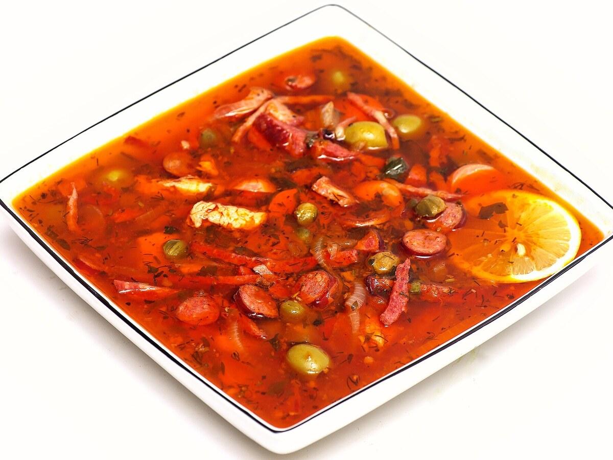 Schüssel rote Suppe mit Wurstscheiben, Oliven, Zitrone und Kräutern.