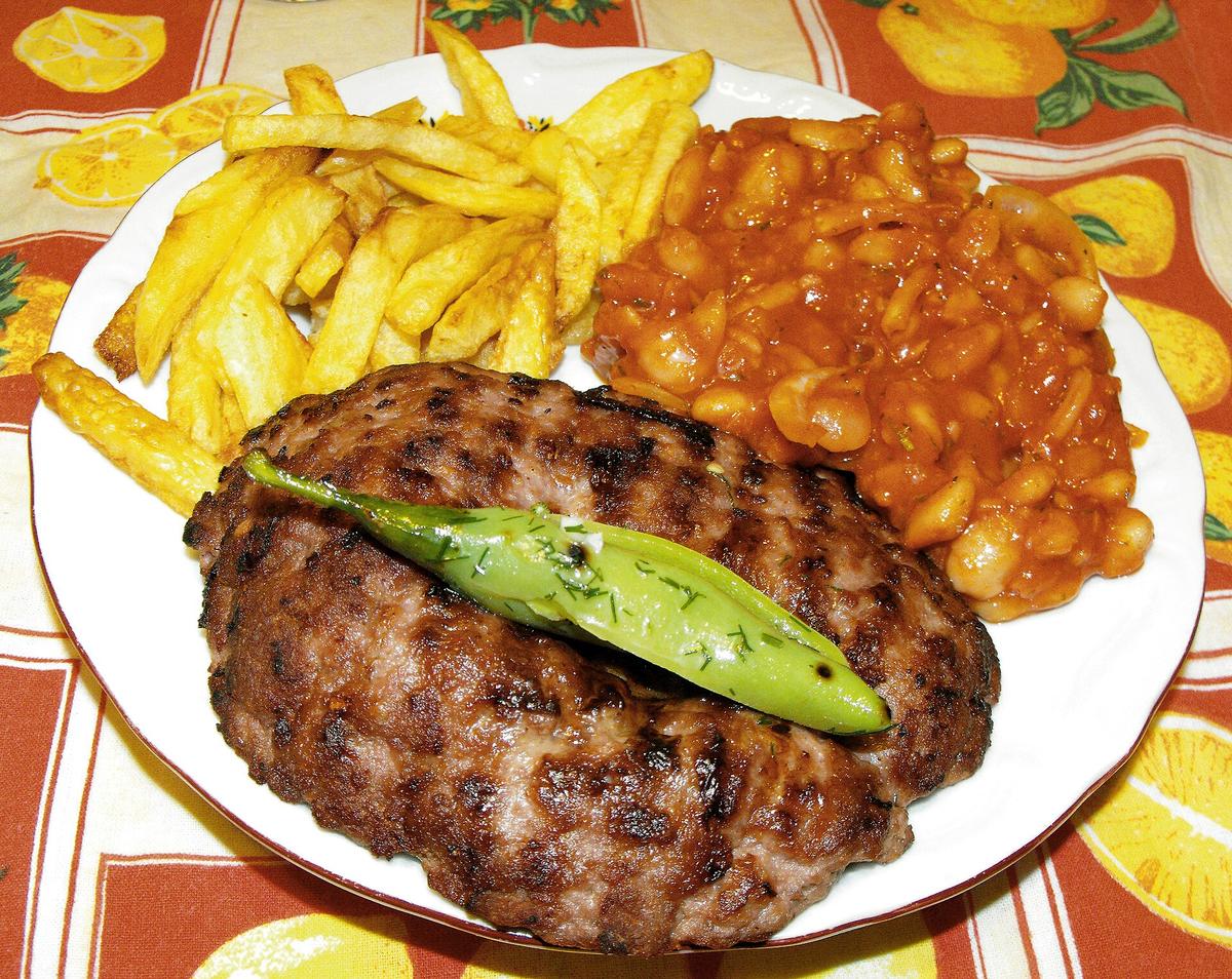 Große gegrillte Hackfleischpastete auf einem Teller mit Pommes Frites, Bohnen und grüner Paprika.