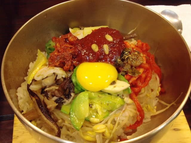 Metal bowl of rice topped with vegetables, beef, red sauce, and an egg.