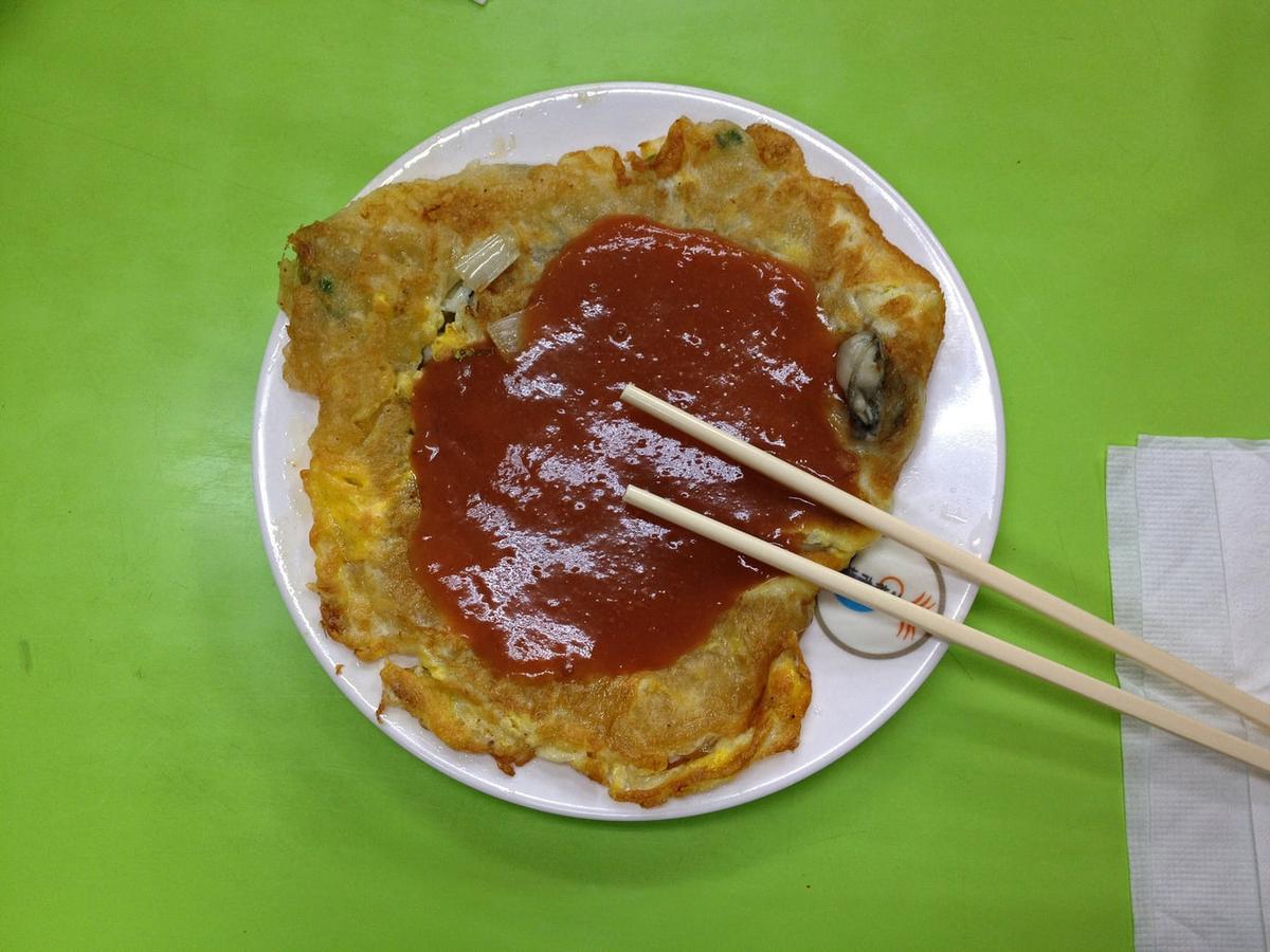 Une omelette aux huîtres avec des légumes verts et une sauce rouge servie sur une assiette blanche.