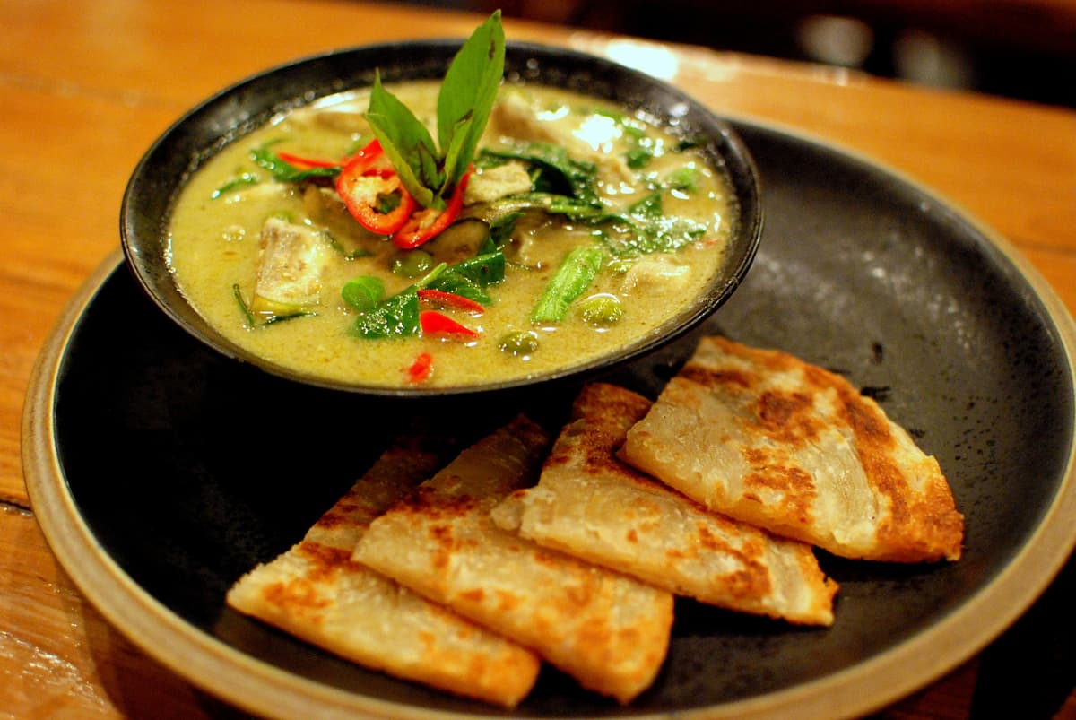 Bowl of pale green chicken curry with herbs and chile beside folded flatbread.