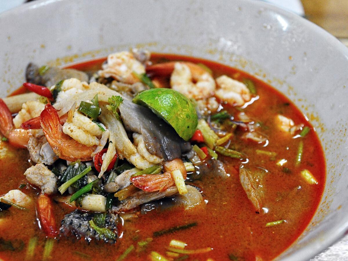 Semangkuk sup udang asam panas merah dengan seafood, bumbu, dan jeruk nipis.