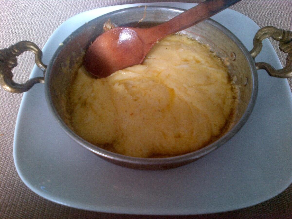 Kleine Metallpfanne mit glänzend geschmolzenem Käse und Maismehl, dazu ein Holzlöffel.