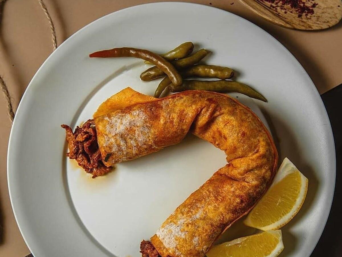Curved meat-filled wrap on a white plate with lemon wedges and green peppers.