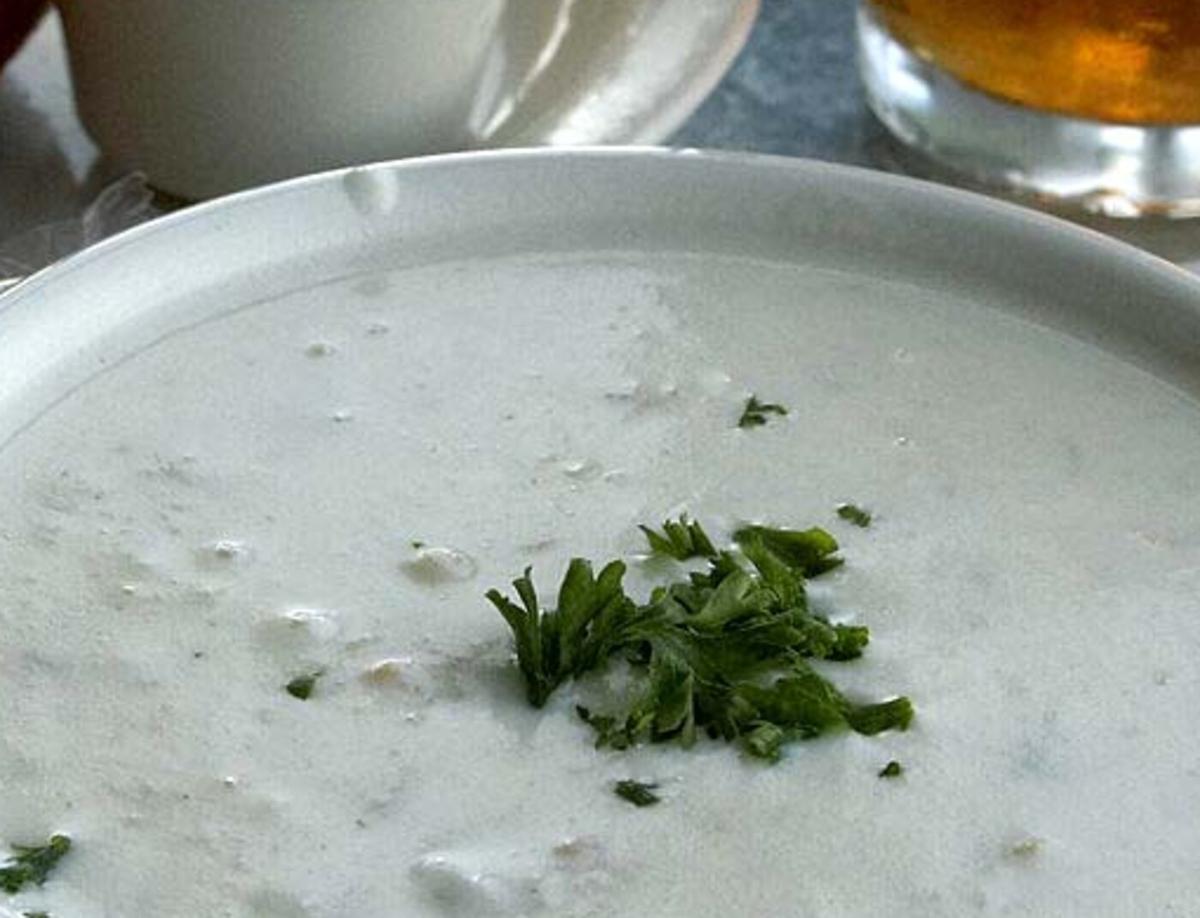Nahaufnahme einer Schüssel mit cremiger Muschelsuppe mit Petersiliengarnitur, Brot und einem Getränk im Hintergrund.