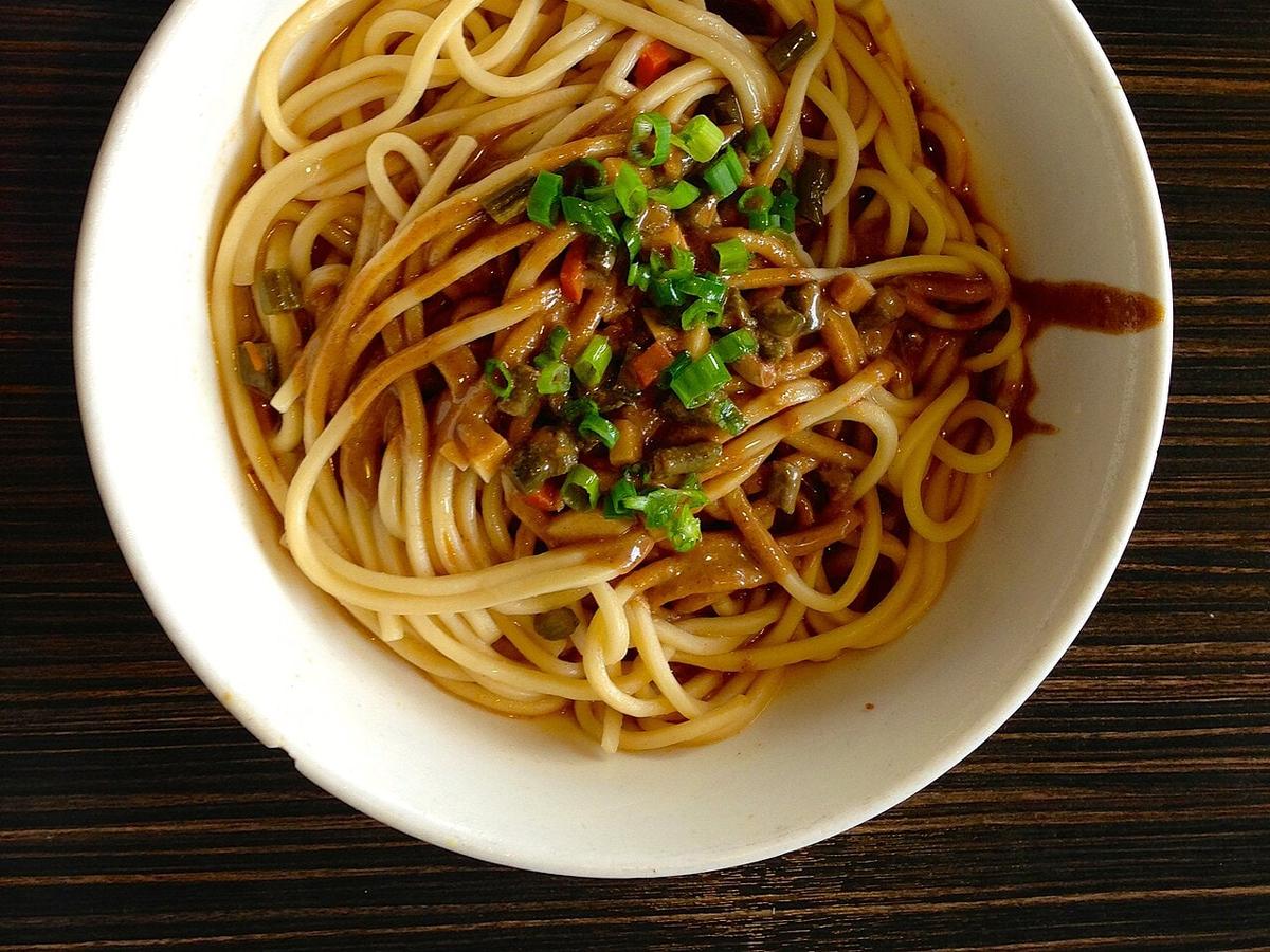 Mangkok mie gandum berlapis saus dengan daun bawang di atas meja kayu gelap.