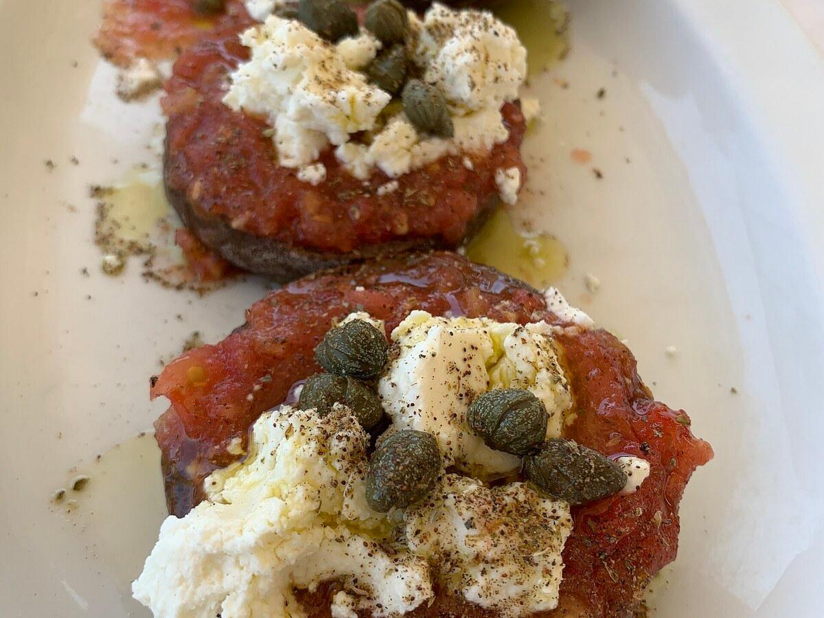 Gerstenröggel belegt mit Tomate, weißem Käse, Kapern, Olivenöl und Kräutern.