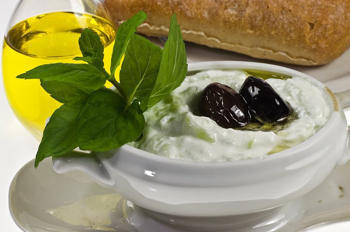 Schüssel mit Gurken-Joghurt-Dip mit Kräutern, Oliven, Olivenöl und Brot.