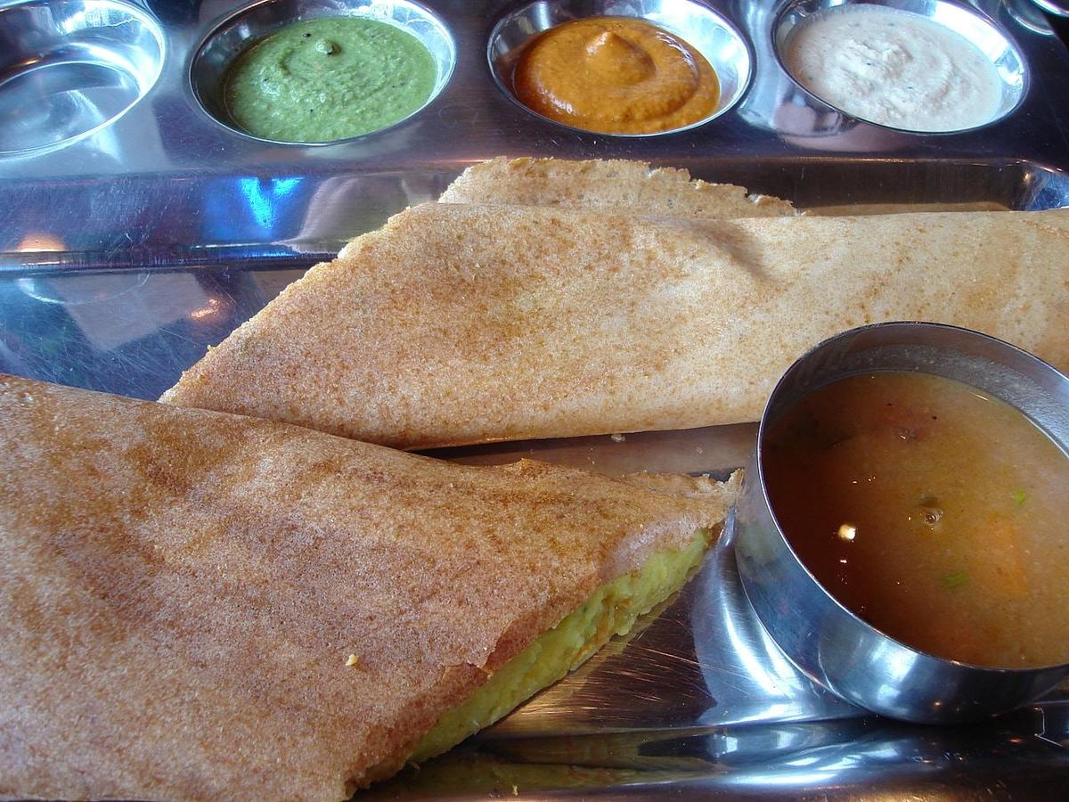 Knusprig gefaltete Dosa auf einem Stahltablett mit Chutneys, Sambar und sichtbarer Kartoffelfüllung an einem Ende.