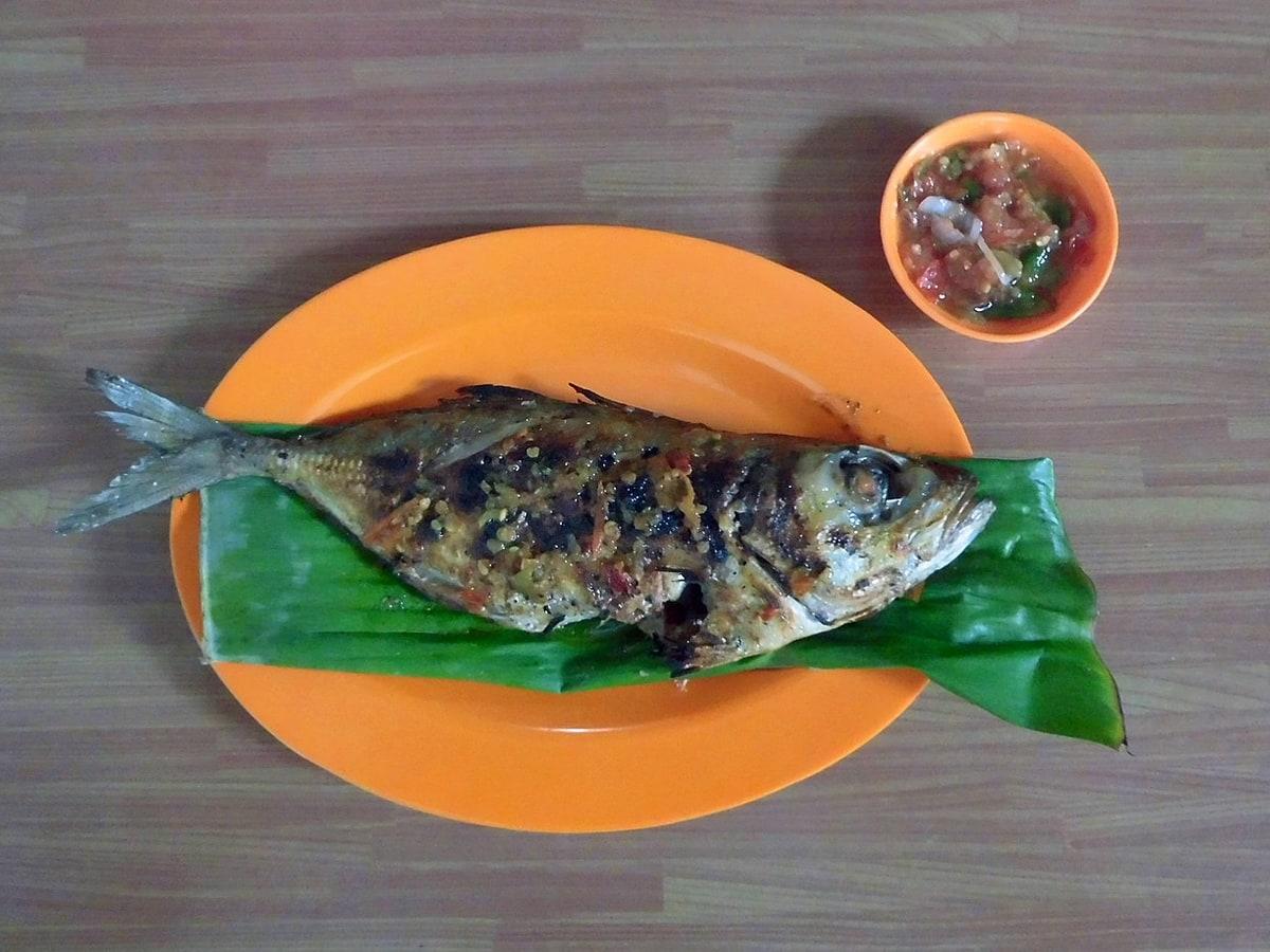 Pescado entero a la parrilla sobre hoja de plátano con un pequeño cuenco de salsa picante roja.