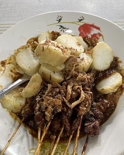 Chicken skewers in peanut sauce with rice cakes and fried shallots on a plate.
