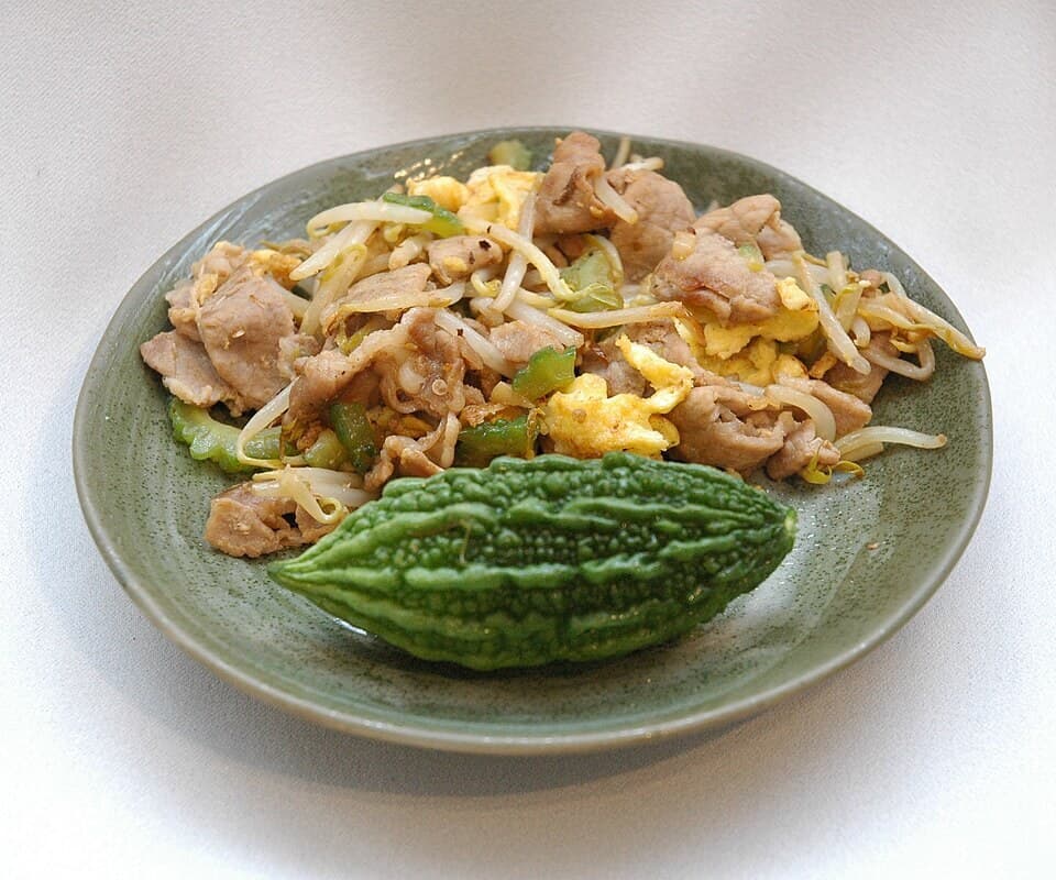 Teller mit Bittermelonenpfanne, Tofu, Ei, Schweinefleisch, Sprossen und halbierte Bittermelone.