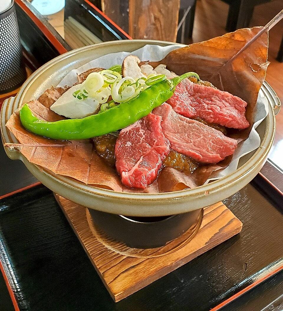 Slices of beef and vegetables cooking over miso on a broad leaf in a small tabletop pan.