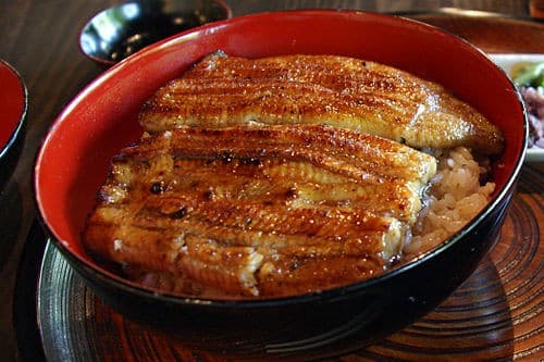 Glossy grilled eel laid over rice in a red bowl on a restaurant table.