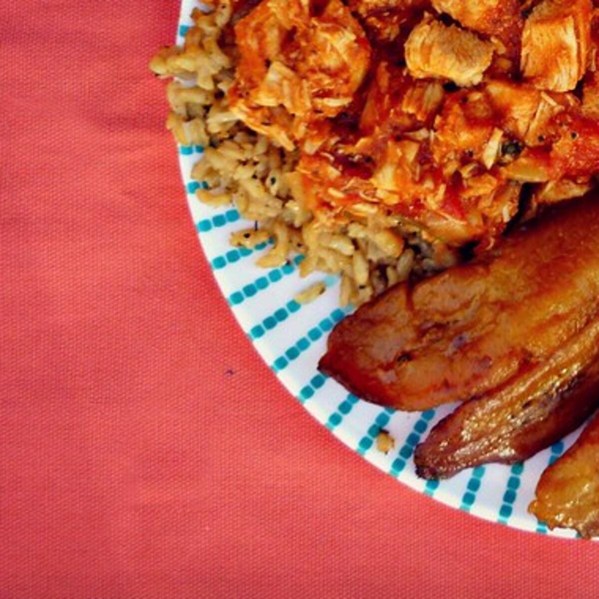 Nahaufnahme von gewürztem Huhn und Reis mit gebratenem Kochbananen auf einem Teller.