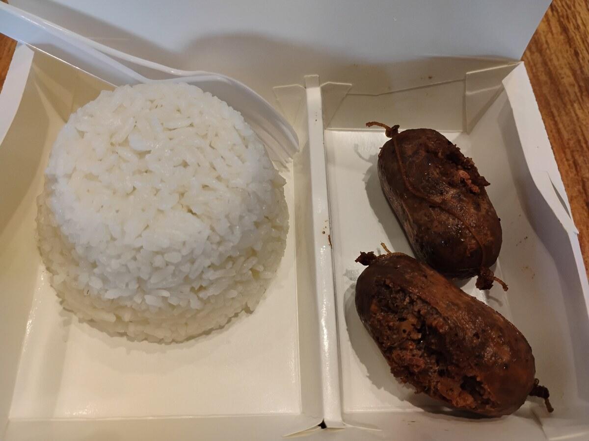 Kleine braune Würste mit Reis in einer schlichten Takeaway-Box serviert.