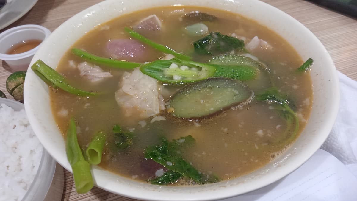 Glass bowl of sour pork soup with ribs, greens, taro, tomato, and long green chiles.