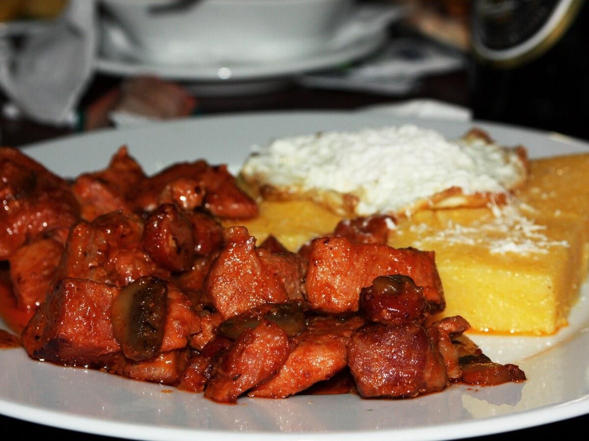 Teller mit gebräunten Schweinefleischstücken, Polenta, Käse, Ei und Sauce.