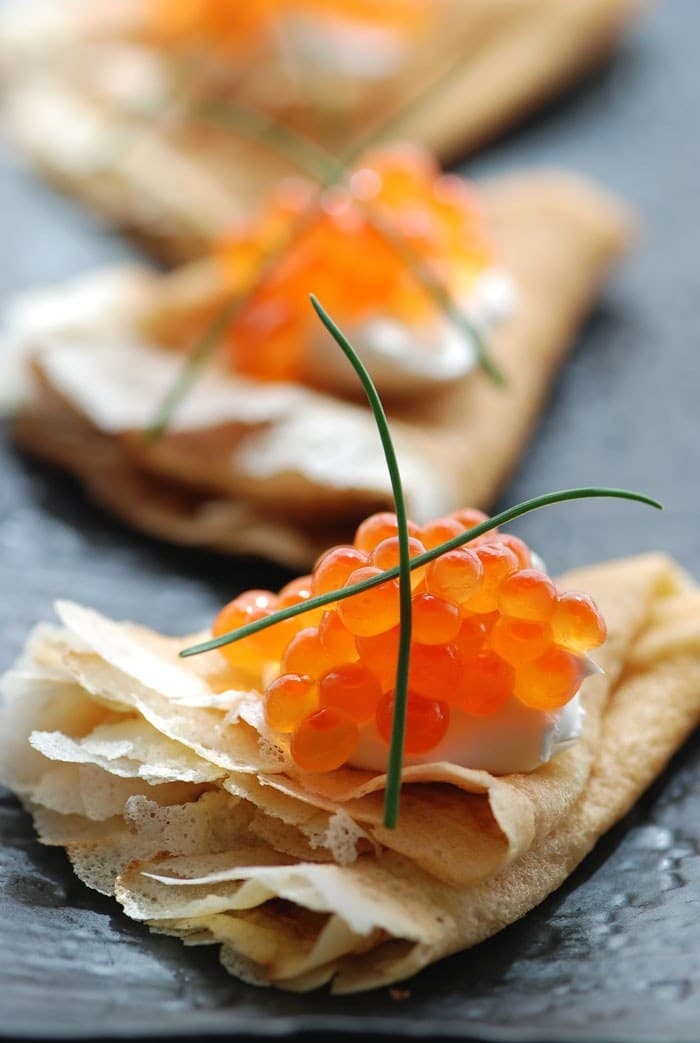 Pancake tipis dilipat dengan topping krim asam, telur ikan oranye, dan hiasan hijau kecil.