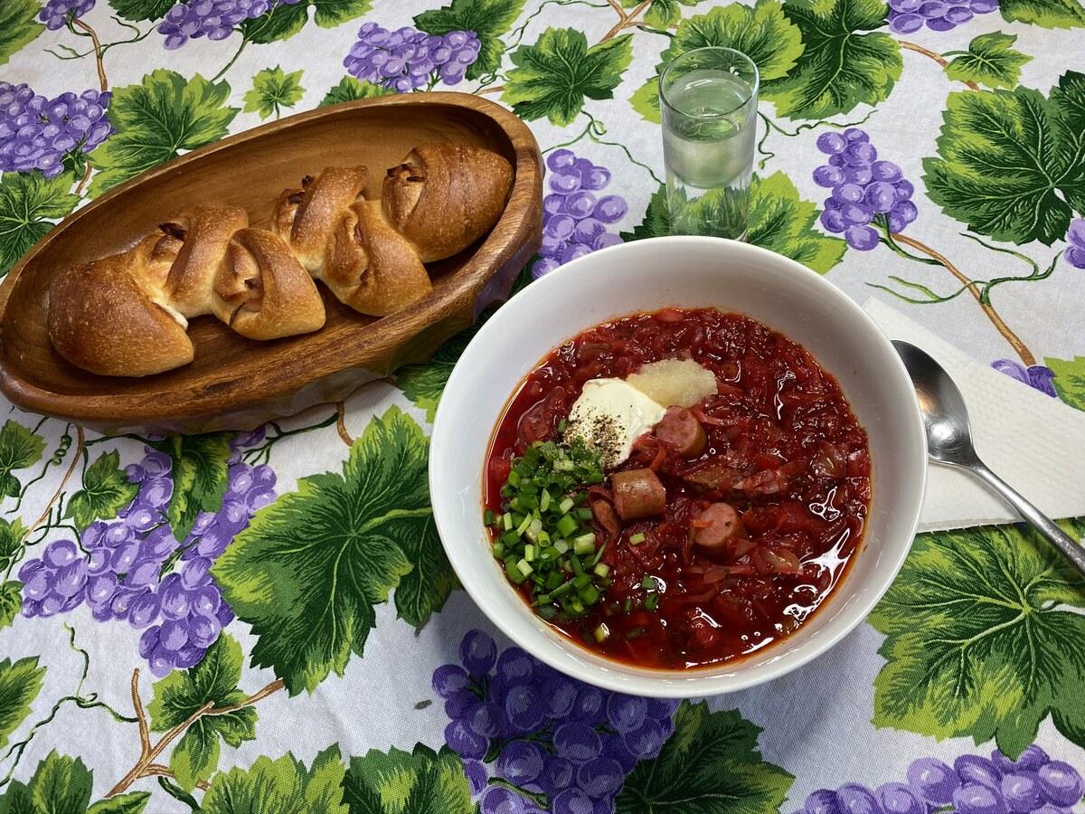 Bowl of red beet soup with sour cream, herbs, and small buns nearby.