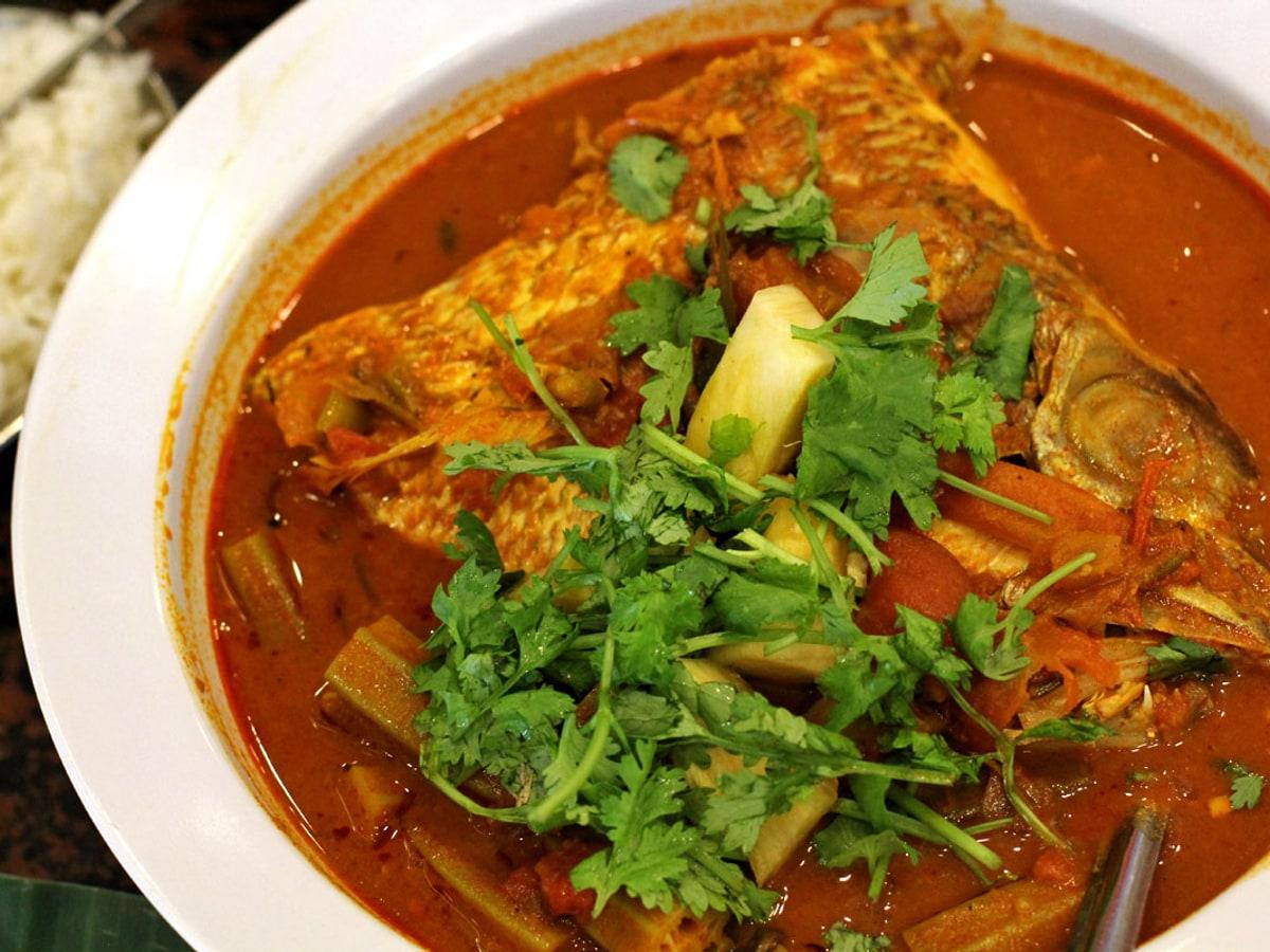 Deep bowl of red-orange fish head curry with herbs and vegetables.
