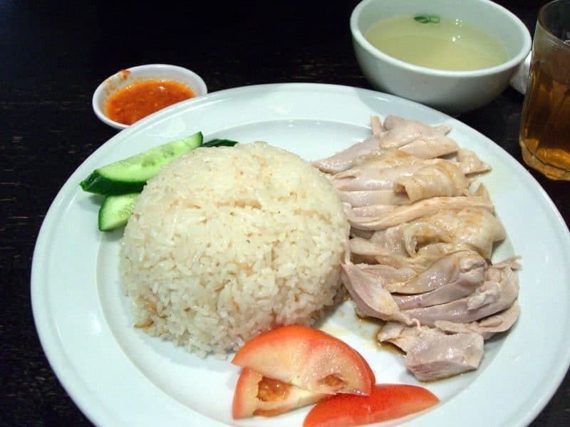 Pollo escalfado en rodajas junto a un montículo de arroz, pepino, salsa y sopa.