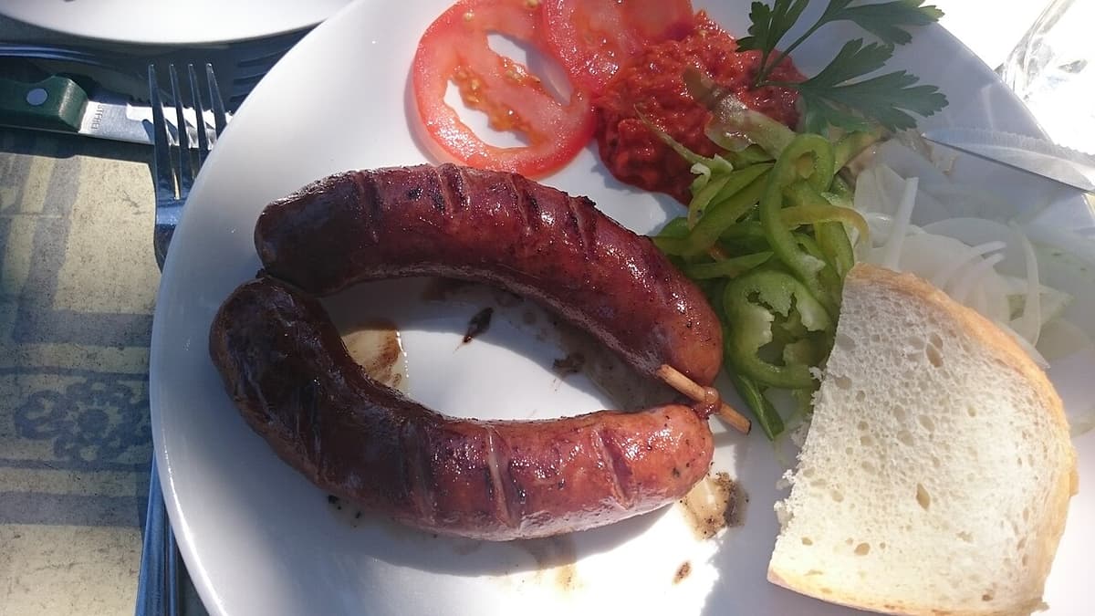 Geräucherte rötliche Wurst auf einem Teller mit Brot, Tomate und grüner Paprika.