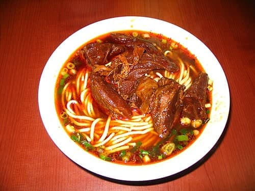 Bowl of dark beef noodle soup with wheat noodles, beef pieces, scallions, and broth.