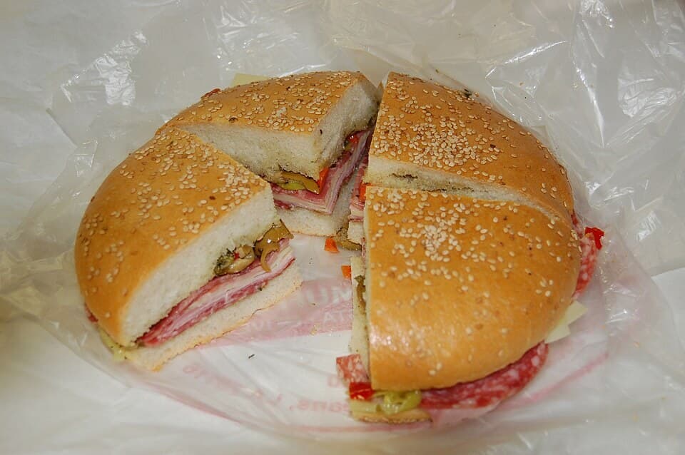 Sándwich redondo con sésamo cortado en secciones, con carnes curadas, queso y ensalada de aceitunas visibles.