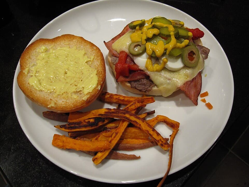 Sándwich cargado de carne con huevo, queso, lechuga, tomate y papas fritas en un plato blanco.